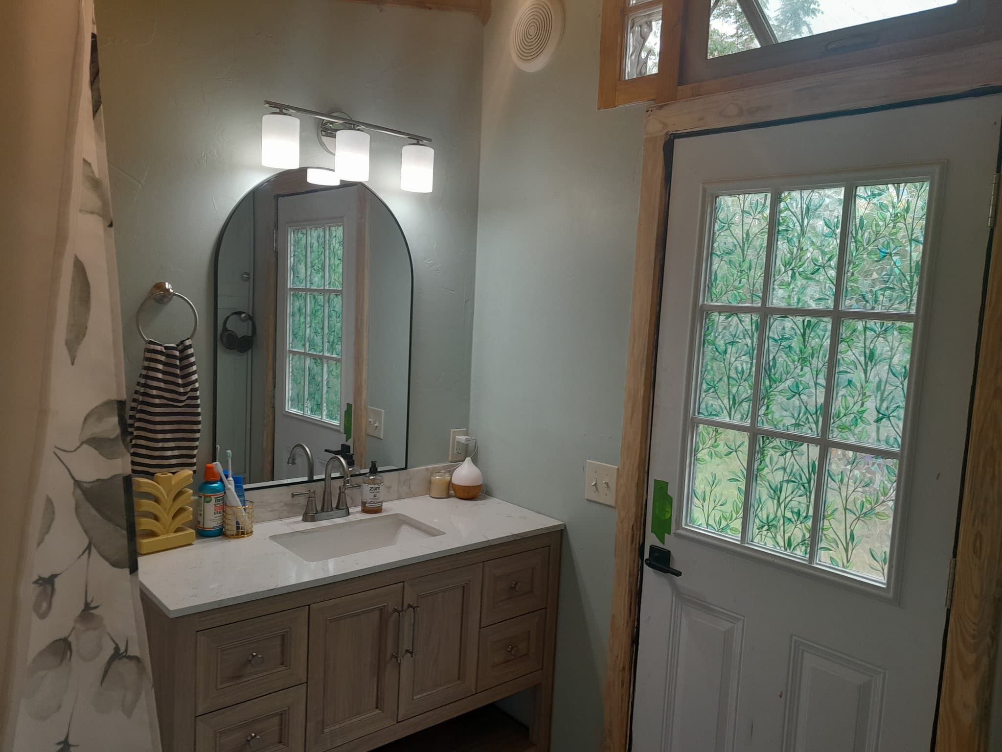 The detached bath house — marble vanity, arched mirror, sage walls, and a leaf-patterned glass door